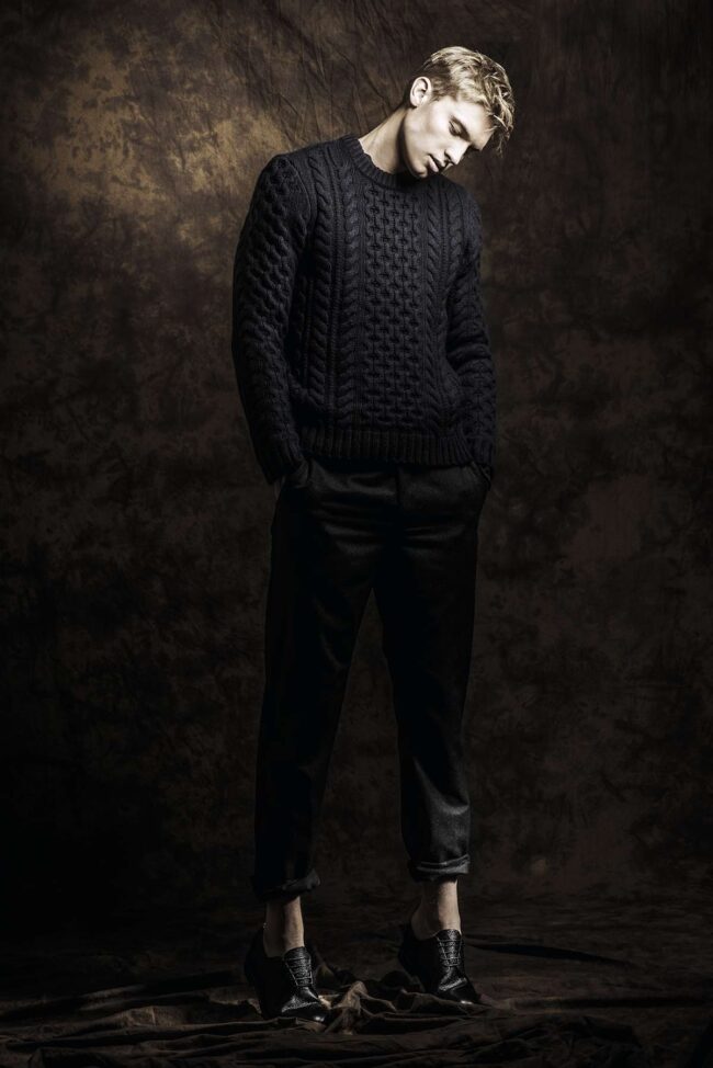Los Angeles fashion photographer, studio menswear fashion image of a male model in a black textured knit sweater and cropped trousers against a dark backdrop, editorial photo for fashion brands, campaigns, lookbooks, and digital marketing.