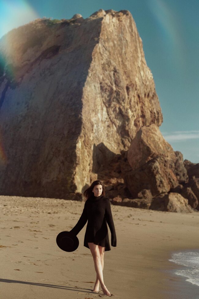Los Angeles fashion photographer, coastal editorial fashion image of a model in a black knit dress walking barefoot along the beach with large rock formations in the background, campaign photo for fashion brands, lookbooks, and digital marketing.