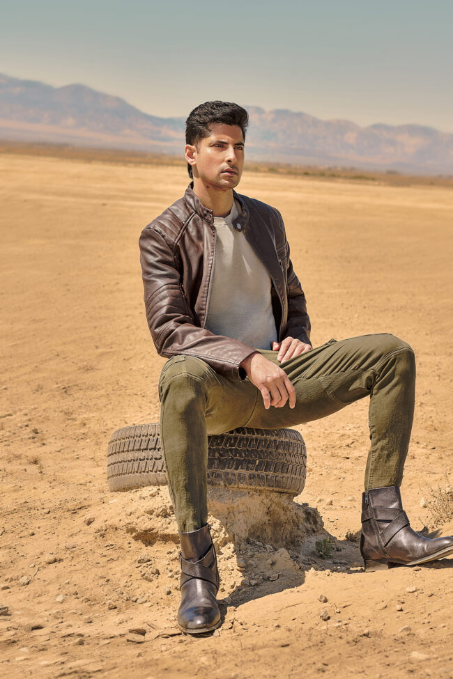 Los Angeles fashion photographer, desert menswear fashion image of a male model in a leather jacket, green pants, and boots seated on a tire with mountains in the background, campaign photo for fashion brands, lookbooks, and digital marketing.