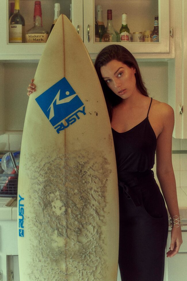 Los Angeles fashion photographer, lifestyle editorial fashion image of a model in a black dress standing with a surfboard in a kitchen, campaign photo for fashion brands, lookbooks, and digital marketing.